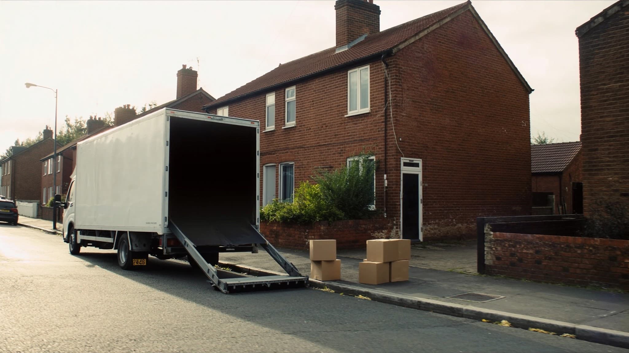 A removals lorry with its tailgate down outside a brick house, boxes on the pavement