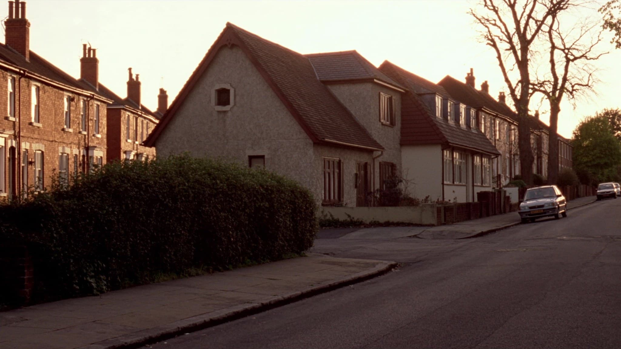 A quiet residential street at golden hour, a rendered house with a tiled roof set back behind a tall hedge, parked cars further down