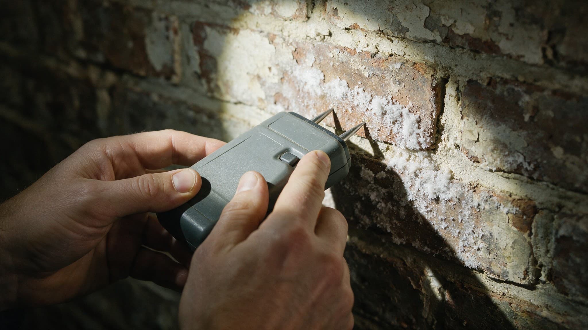 A hand holding a damp meter against a bare brick wall with salt staining