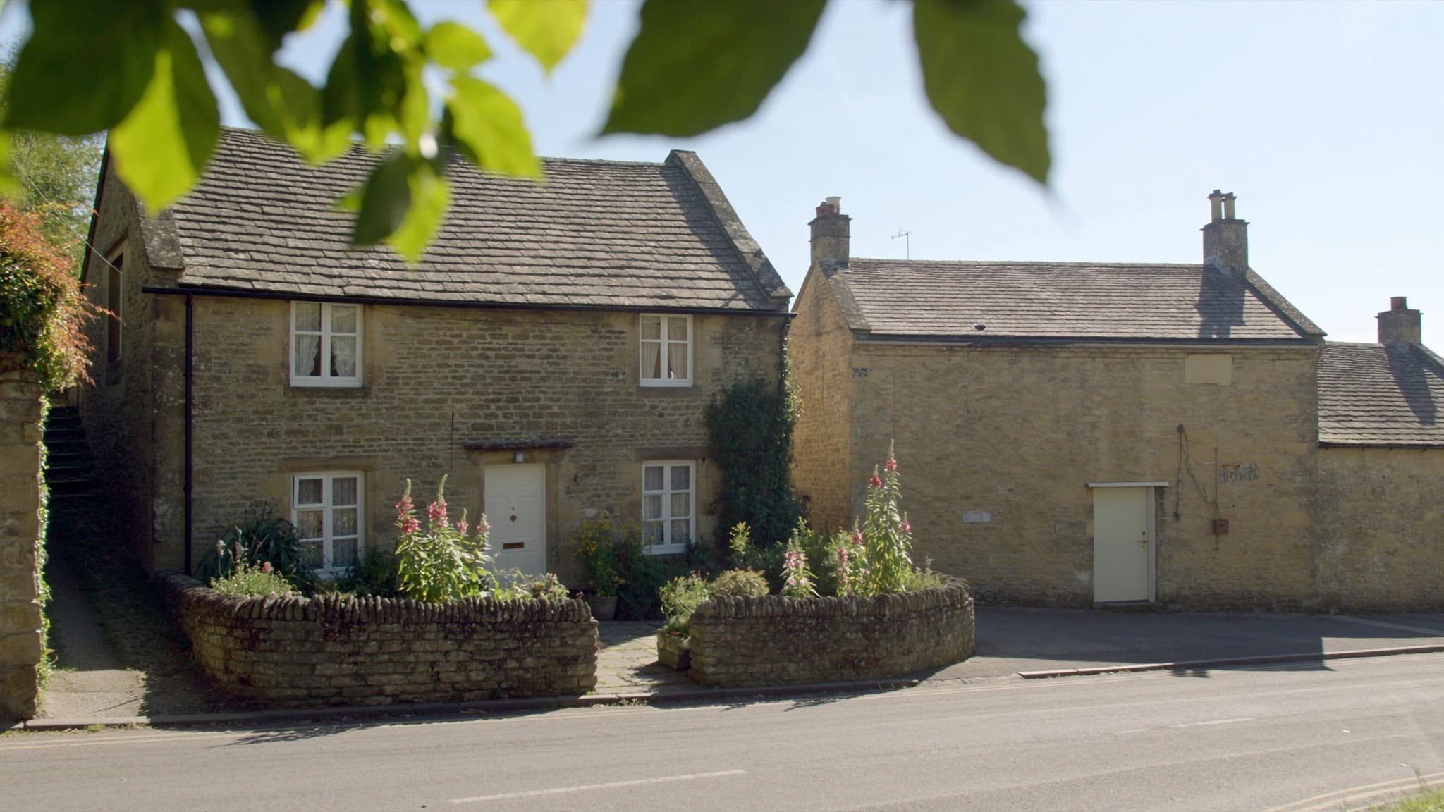A honey coloured stone cottage on a village lane with flowers in the front beds