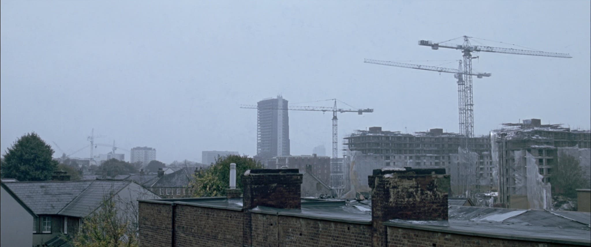 Cranes and half-finished rooftops on a city edge under a flat grey sky.