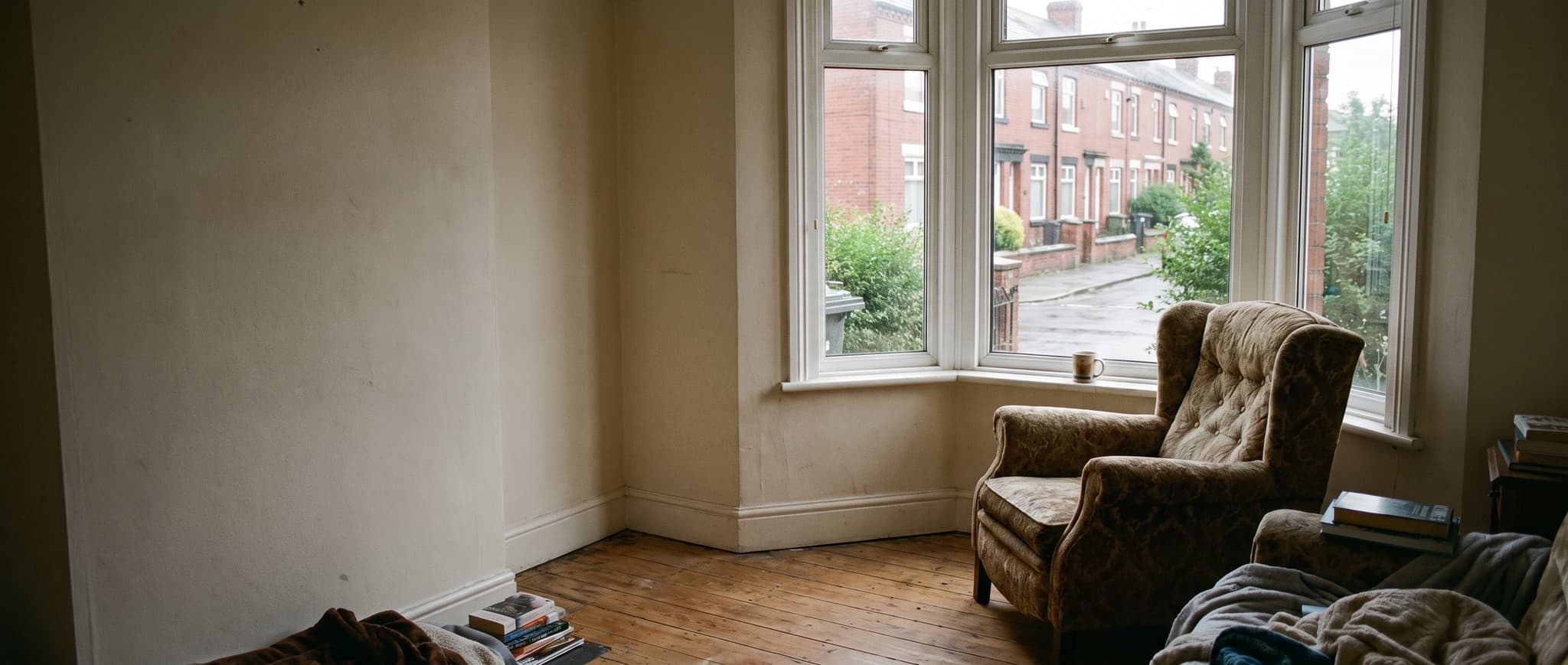 An armchair beside a bay window in a British living room, a mug on the sill and a terraced street outside