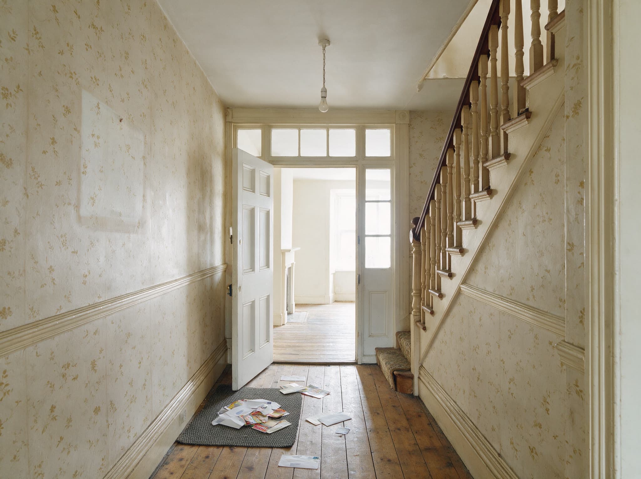 The hallway of an empty British terraced house, unopened post scattered across the doormat and bare floorboards, a door standing open onto an empty room