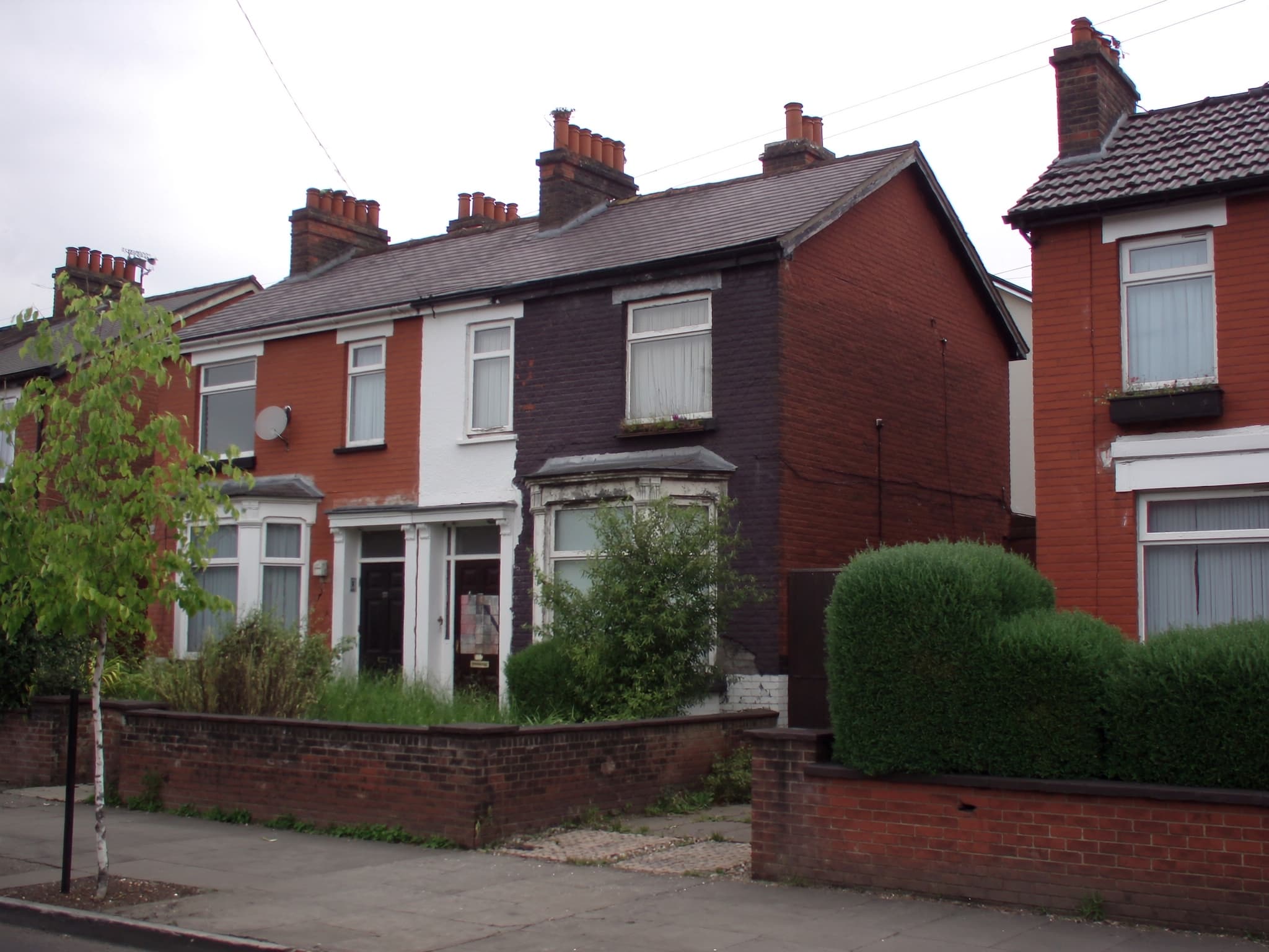 A neglected end-of-terrace house with stained brickwork and an overgrown front garden, standing between cared-for neighbours with clipped hedges