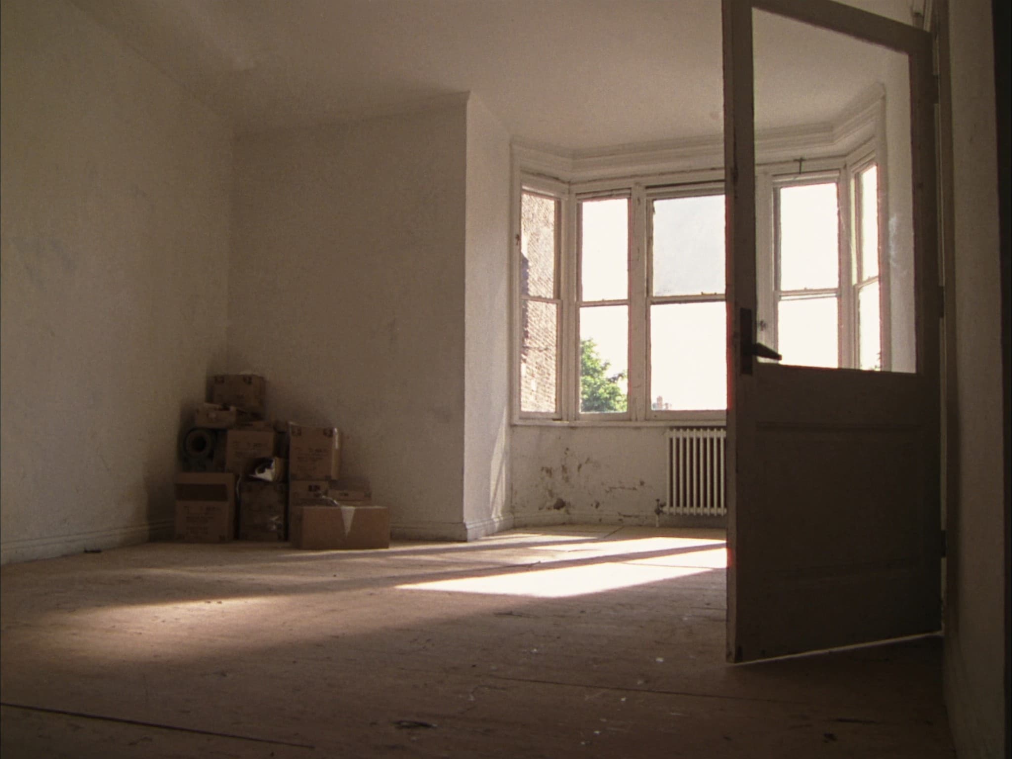 An empty living room in a British terraced house on moving-in day, sunlight across bare floorboards and unopened boxes stacked against the wall