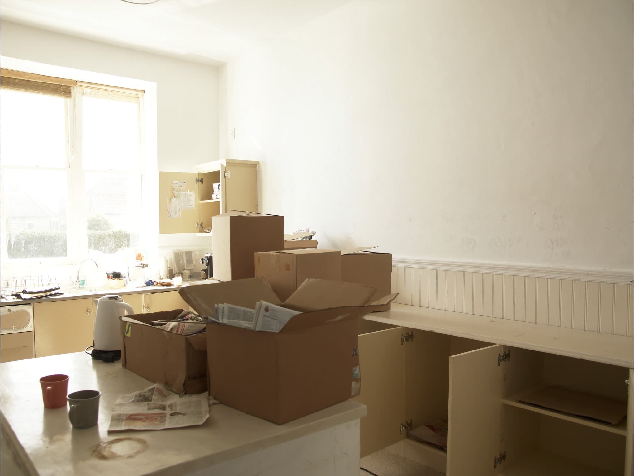 A British kitchen mid-move, packing boxes along the worktop, a kettle and two mugs beside them and cupboards standing open