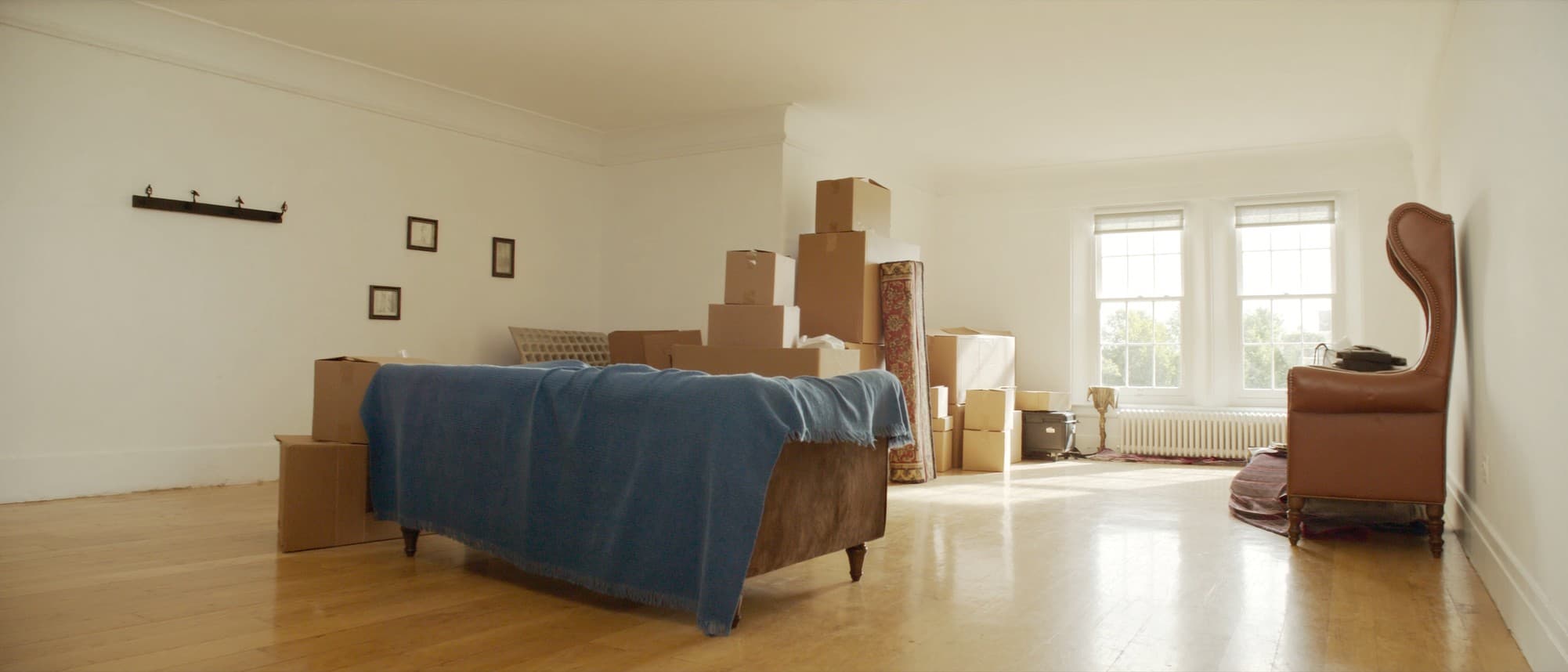 A bright British living room halfway through a move, cardboard boxes stacked along one side, a sofa pulled away from the wall and pictures taken down