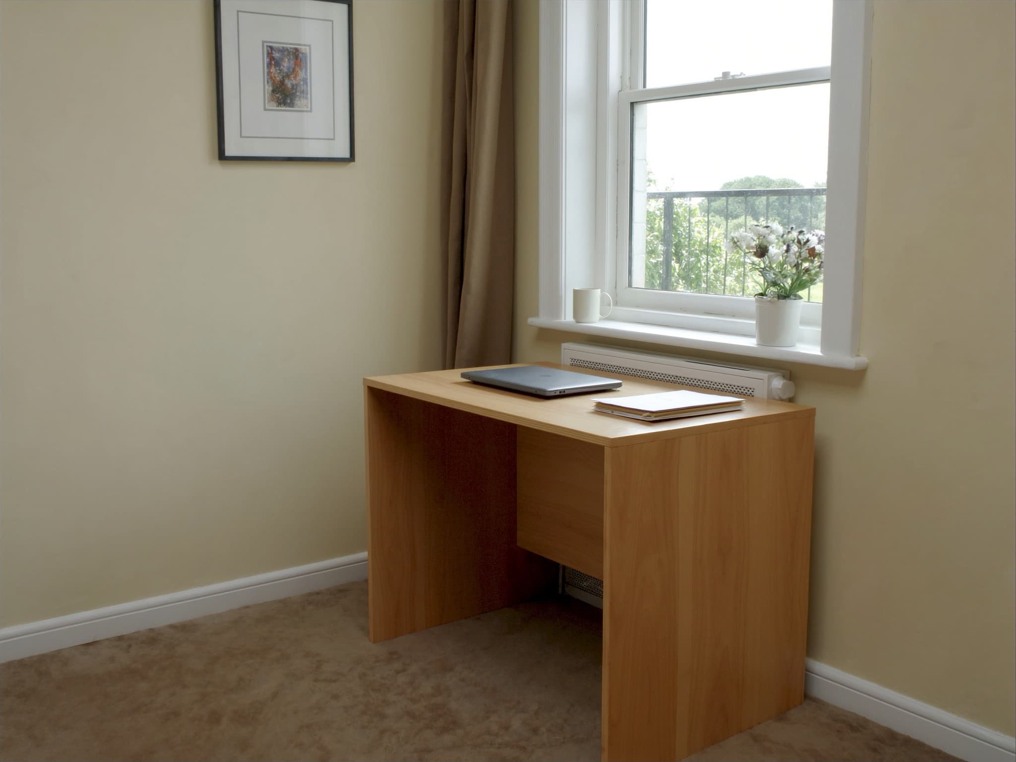 A corner of a British spare room used as a home office, a plain desk under a sash window with a closed laptop and a stack of letters
