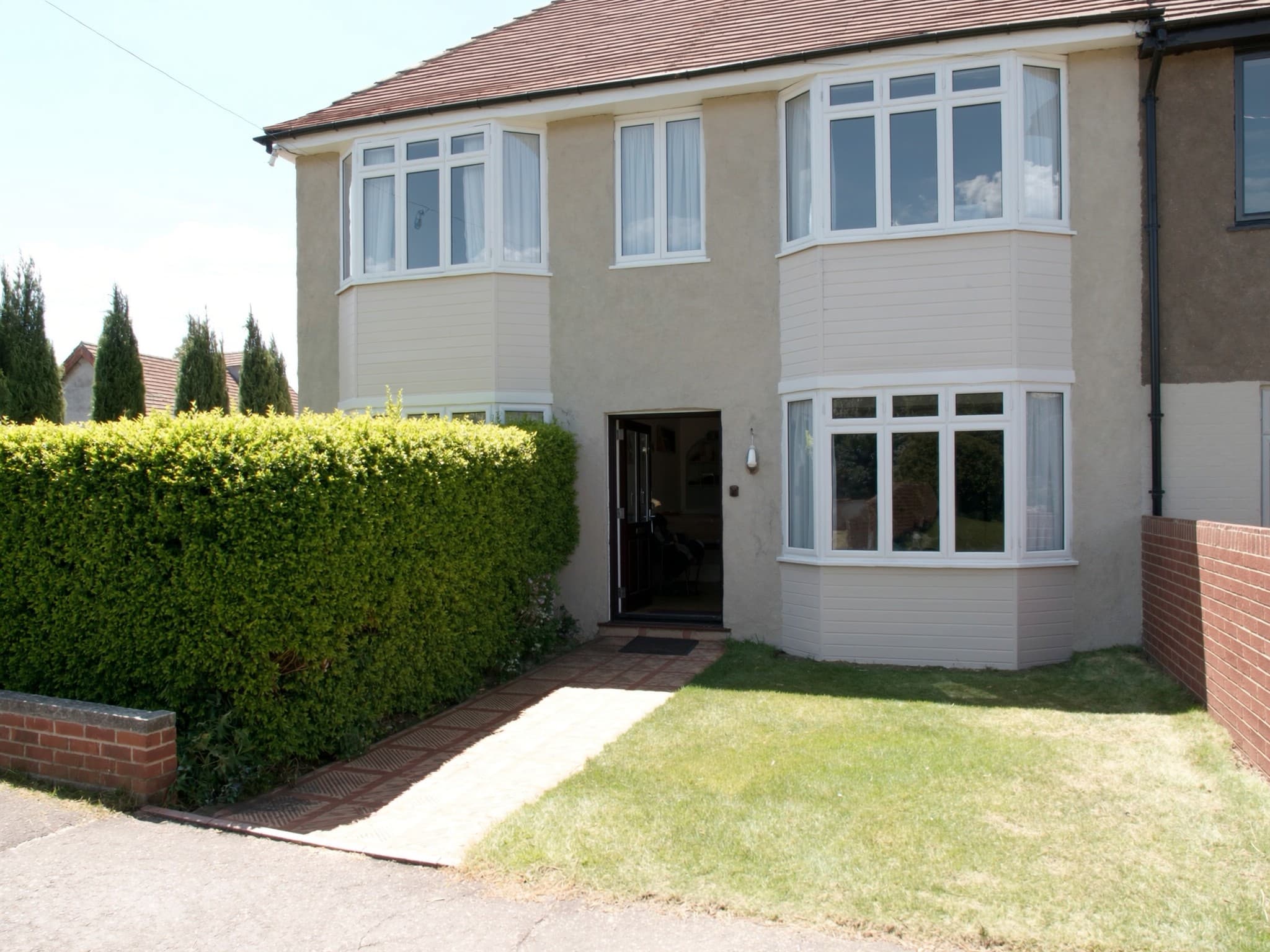 A British bay-fronted house on a bright morning with the front door open, a clipped hedge and a freshly mown front lawn