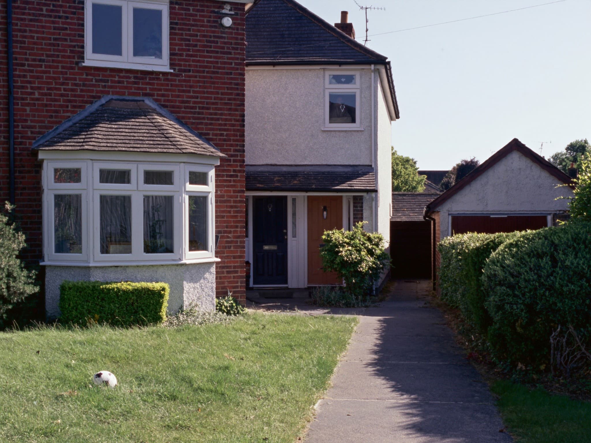 An ordinary British semi-detached family home seen from the front garden path, a bay window, a panelled front door and a football on the lawn