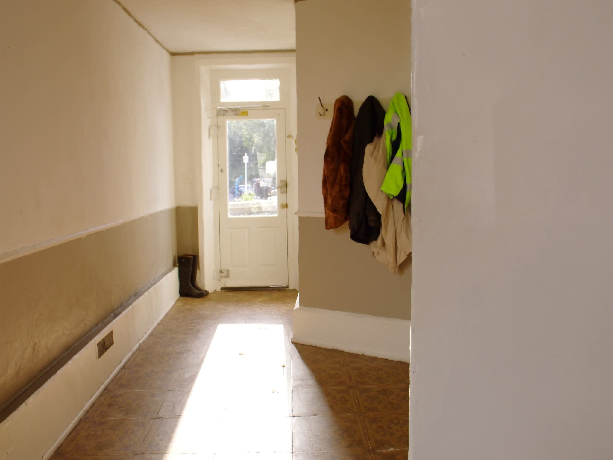 A British hallway with a row of coat hooks, several coats hanging and one hook left empty, sunlight across patterned floor tiles