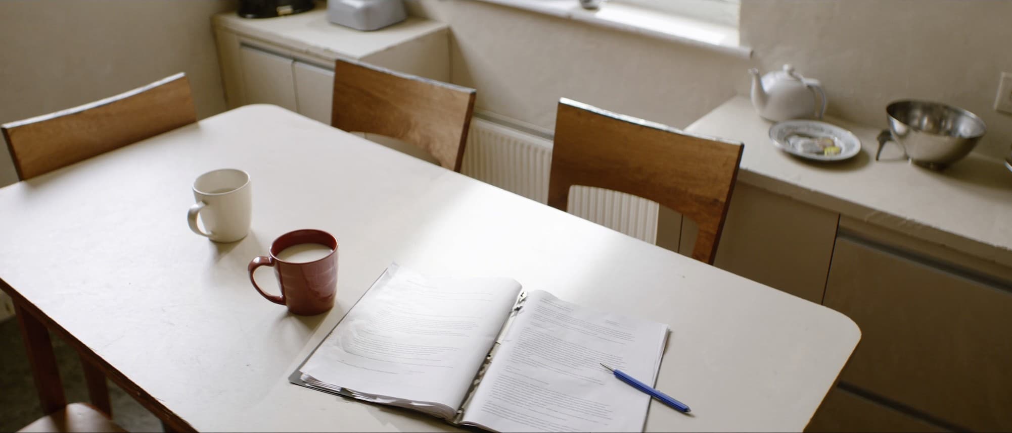 A bright British kitchen table with two chairs pulled up on the same side, an open folder of paperwork lying between two mugs of tea