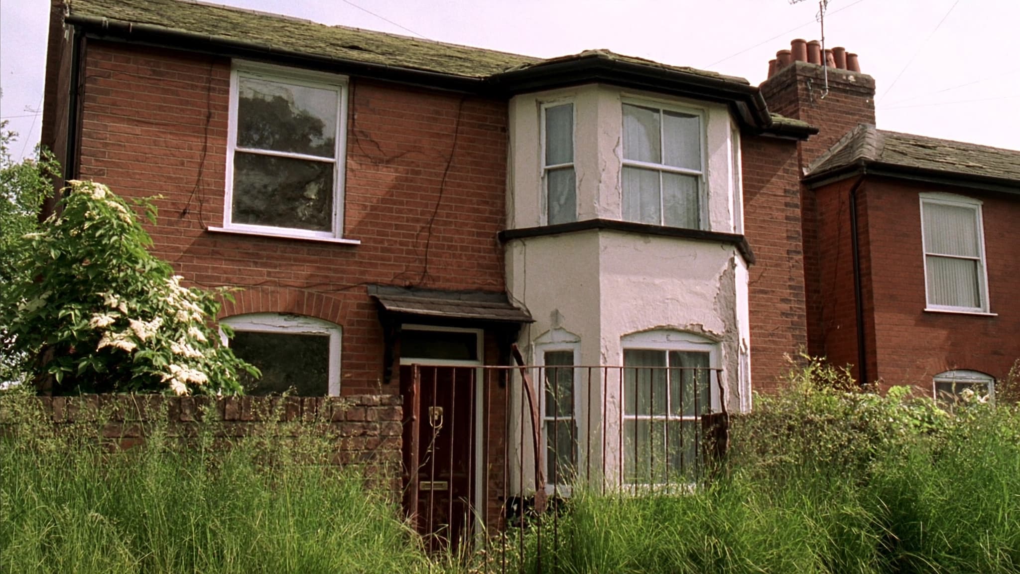A tired 1930s bay-fronted semi with cracked render, peeling paint and an overgrown front garden behind rusted railings