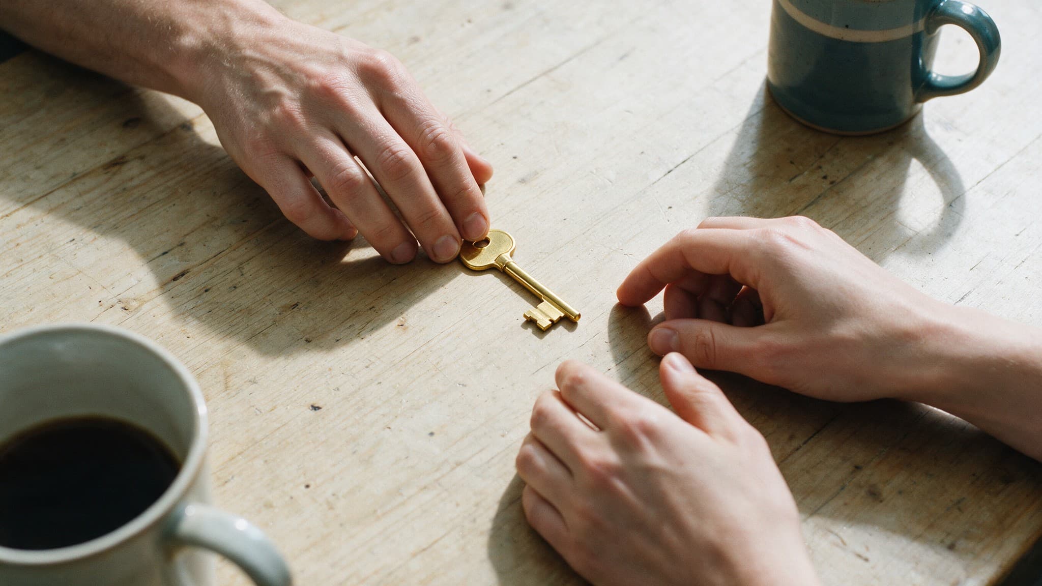 One pair of hands passing a single house key to another across a kitchen table
