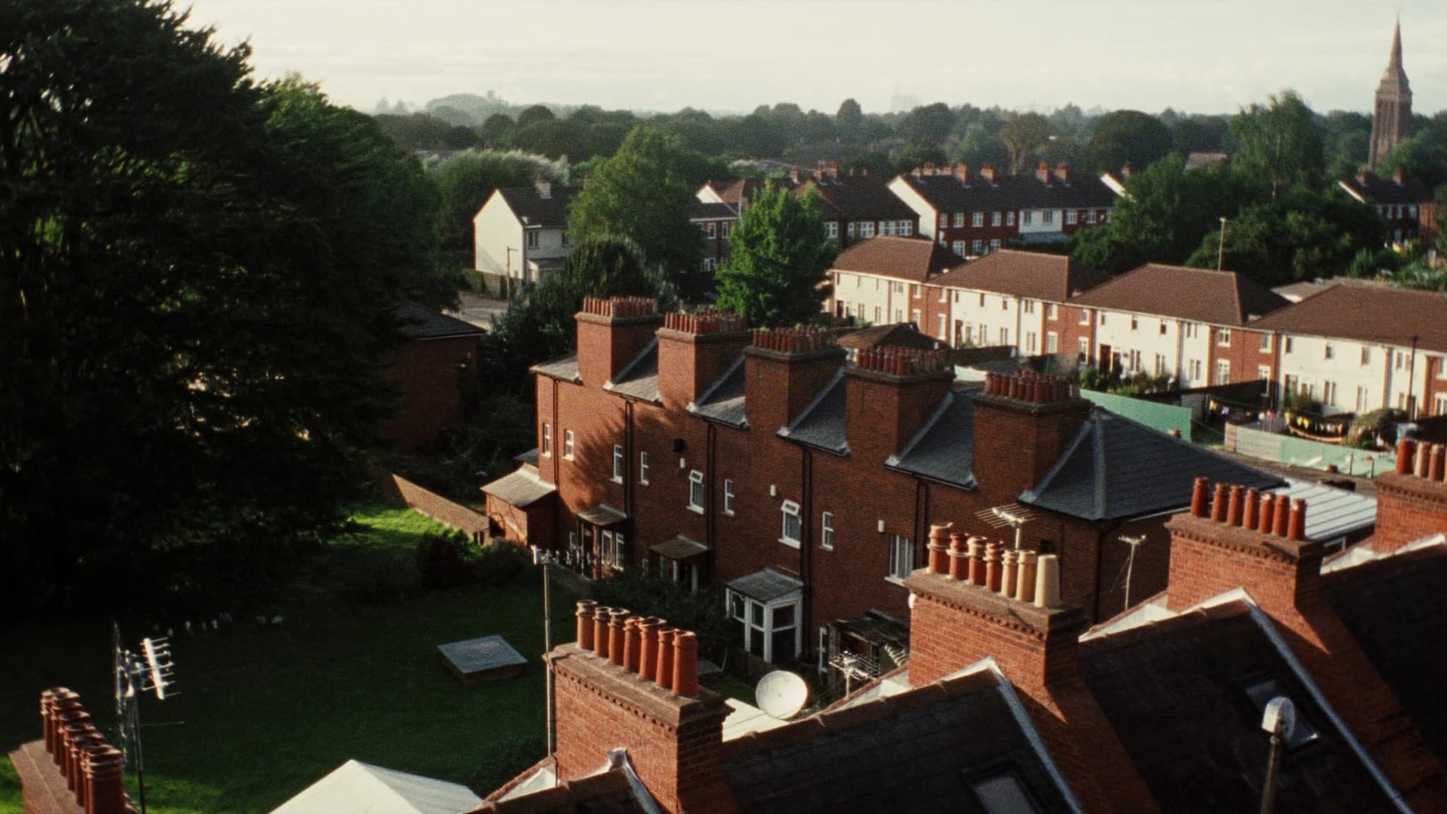 An elevated view across a British neighbourhood in evening light, red-brick terraces with chimney pots and long back gardens, rendered houses and a church spire beyond